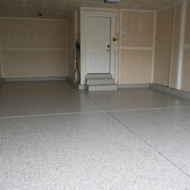 An empty garage with light beige walls and a gray speckled epoxy floor, leading to a white door with steps.
